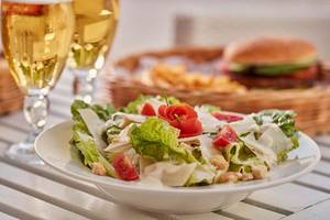 Ein Teller mit einer Salatmischung und einer Rose, mit zwei vollen Weingläsern und einem Korb mit Pommes und Burgern im Hintergrund.
