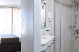 Badezimmer im Kriti Beach Hotel mit Dusche, Waschbecken und Bett im Hintergrund.