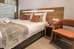 Schlafzimmer im Adele Beach Hotel mit Doppelbett, Dekokissen, Nachttisch, Stuhl und Eingangstür.
