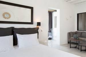 Ein modernes Hotelzimmer von Kivotos Mykonos mit weißem Bett, grauem Sofa, Tisch und Uhrspiegel.