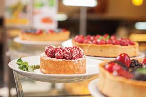 Mehrere Desserts, darunter eine Tarte mit Himbeeren und eine mit schwarzen Beeren, präsentiert auf weißen Tellern in einem Glasschrank.