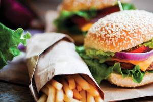 Zwei Burger und Pommes auf braunem Papier, eines mit Zwiebeln und Salat, das andere mit Tomaten und Salat.