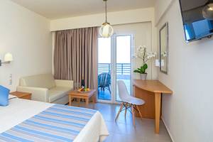 Innenansicht eines Hotelzimmers im Themis Beach Hotel mit Bett, Schreibtisch und Balkon.