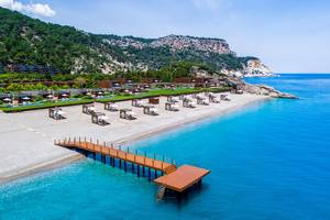 Luftaufnahme des Maxx Royal Kemer Resorts mit weißen Sonnenliegen auf dem Strand und blauem Meer.