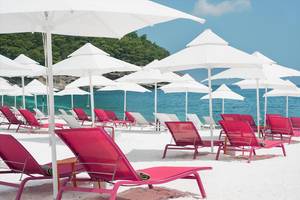 Strand des Maxx Royal Kemer Resorts mit roten Liegestühlen und weißen Sonnenschirmen.