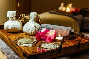 Ein Holztablett mit Spa-Zubehör im Crystal Springs Beach Hotel, einschließlich zweier weißer Beutel, einem Handtuch, Blumen, Kerzen und kleinen Glasbehältern.