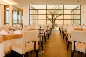 Innenansicht eines Restaurants im Astir Odysseus Kos Resort & Spa mit gedeckten Tischen und Blick auf einen Olivenbaum.