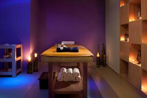 Massageraum im Blue Bay Resort mit violetter Wand, Massageliege, gestapelten Handtüchern und dekorativen Kerzen.
