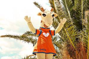 Maskottchen des Ostria Resort & Spa vor einer Palme, das ein orangefarbenes Outfit mit dem Wort Lollo trägt.
