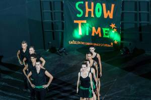 Tanzgruppe auf der Bühne unter einem 'Show Time'-Banner, das mit grünem Licht beleuchtet ist.
