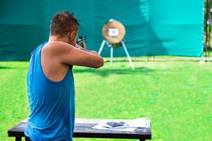 Ein Mann in einem blauen Tanktop zielt mit einer Waffe auf eine Zielscheibe auf einer Wiese.