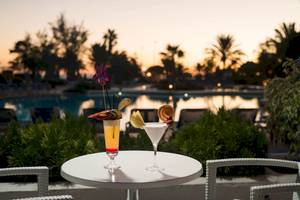 Zwei Cocktails mit Früchten auf einem Tisch mit Blick auf einen Pool und Sonnenuntergang im Hintergrund.