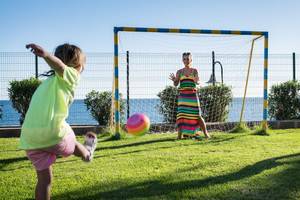 Ein Kind spielt mit seiner Mutter Fußball im Freien, ein Tor im Hintergrund.