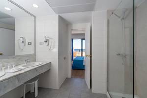 Badezimmer im Grand Teguise Playa mit Doppelwaschbecken, Spiegel, Föhnen und einer offenen Tür zum Schlafzimmer mit einem Bett.