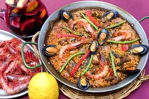 Eine Schüssel Paella mit Garnelen, Muscheln, Tomaten und Paprika auf einem Tisch neben einem Glas Sangria und einer Zitrone.