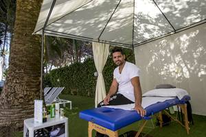 Ein Mann sitzt auf einem Massagestuhl unter einem Zelt im Freien, nahe einer Palme und mehreren Sonnenliegen, im Cosmopolitan Hotel.