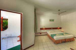 Innenansicht eines Hotelzimmers im Fihalhohi Island Resort mit einem Bett, Decken und Kissen, einem Ventilator an der Decke und einem offenen Türbogen.