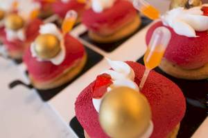 Mehrere rote Desserts mit goldenen Kugeln und orangefarbenen Stäbchen auf einem weißen Teller.