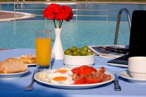 Frühstück am Pool des Plaza Beach Hotels mit rotem Blumenstrauß, Orangensaft, Spiegeleiern, Tomaten, Speck und Laptop.