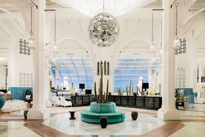 Innenansicht der Lobby des H10 Timanfaya Palace mit zentralem Kaktus in blauem Topf und Spiegel-Pendelleuchte.