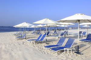 Strandszene mit vielen weißen Sonnenschirmen und blauen Strandliegen im Sand