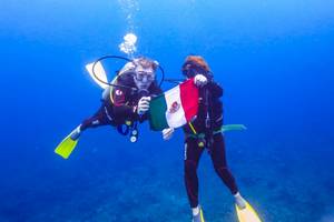 Zwei Taucher im Wasser halten eine mexikanische Flagge vor sich
