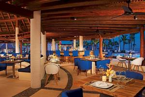 Innenbereich des Restaurants im Secrets Akumal Riviera Maya mit blauen Stühlen und Tischen, Holztischen, Deckenventilatoren und Blick auf den Ozean.