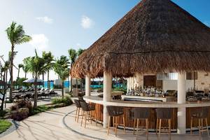 Eine Bar mit Strohdach im Secrets Akumal Riviera Maya Hotel, umgeben von Palmen und Sonnenliegen.