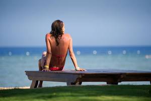 Frau im roten Bikini sitzt auf einer Bank am Strand und blickt auf das Meer.