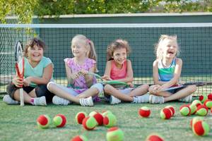 Vier Maedchen sitzen auf einem Tennisplatz, lachen und halten Tennisschlaeger in der Hand, mit Tennisbaellen um sie herum.