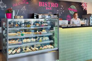 Innenansicht der Bistro Bar im H10 Rubicon Palace mit einer Wandtafel, die 'Bistro Bar' und 'ice cream' beschriftet ist, einer Verkäuferin und einer Vitrine voller Kuchen und Sandwiches.