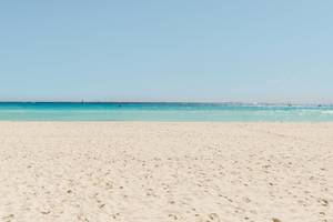 Foto von einem weißen Sandstrand und türkisfarbenem Wasser, mit einem blauen Himmel.