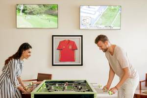 Gäste im Allegro Playacar Hotel spielen Tischfußball vor einem Fußballtrikot und Fernsehern an der Wand.