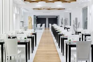 Innenansicht des Restaurants im Dimitra Beach Hotel & Suites mit weißen Tischen und Stühlen, einem hölzernen Gang und Kaktus-Dekorationen.
