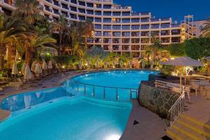 Abendansicht des Swimmingpools im Seaside Sandy Beach Hotel mit illuminierten Gebäuden und Sonnenliegen.
