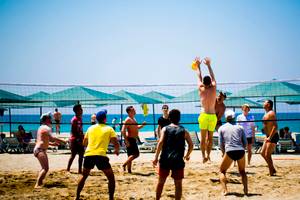 Mehrere Männer spielen Volleyball auf einem Strand mit einem Netz und Sonnenschirmen im Hintergrund.