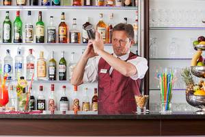 Ein Barkeeper im roten Jackett des Zora Hotels rührt einen Cocktail in einem Glas.