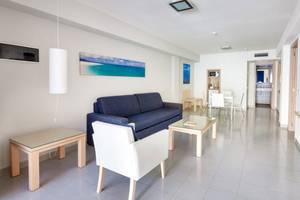 Inneneinrichtung des Tagoro Family & Fun Costa Adeje mit einem blauen Sofa, weißem Stuhl, Glastisch und Fernseher an der Wand.