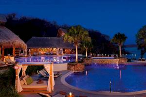 Nachtansicht des Secrets Papagayo Costa Rica Hotels mit beleuchtetem Infinity-Pool und Palmen.