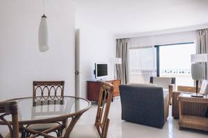Innenansicht eines Hotelzimmers im Iberostar Origin Bella Costa mit Tisch, Stühlen, Fernseher und großem Fenster.