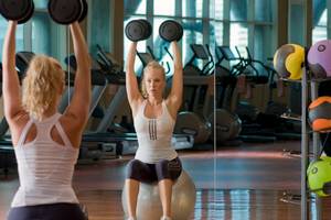Zwei Frauen im Fitnessstudio machen Übungen mit Hanteln auf einem Gymnastikball.