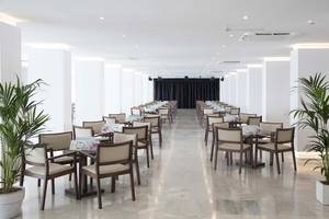 Innenansicht des Speisesaals im Alua Leo Hotel mit geräumigen weißen Wänden, braunen Stühlen und Tischen, sowie grünen Pflanzen in Töpfen.