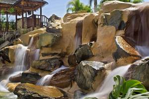 Ein Wasserfall im Tabaiba Princess Hotel mit einer hölzernen Brücke und Pflanzen im Hintergrund.