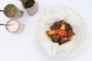 Ein Teller mit gewürztem Fleisch, Tomaten und Kartoffeln, alles in weißem Papier eingewickelt, neben zwei Stoffbeuteln mit Gewürzen.