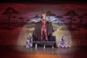 Schauspieler in roter Kleidung als König der Löwen auf einer Bühne mit zwei sitzenden Schauspielern im Hintergrund.