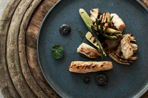 Ein Gericht auf einem blauen Teller mit Holzuntergrund, bestehend aus gegrilltem Fisch, Gemüse und schwarzen Soßen.