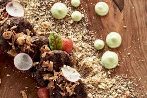 Essenplatte mit schwarzen Burgern, Radieschen, Tomaten, grünen Saucen und Krusten auf einem Holztisch.
