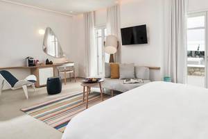 Innenansicht eines Zimmers im Myconian Kyma Hotel mit großem Bett, Couch, Tisch und Fenstern.