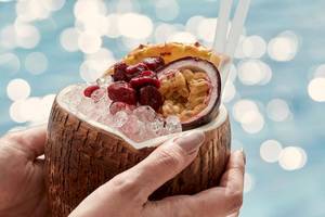 Cocktail im Kokosnuss-Becher mit Früchten und Eis, gehalten von Händen neben einem Pool im Myconian Kyma Hotel