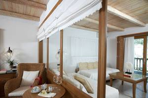 Innenansicht eines Schlafzimmers im Mitsis Blue Domes Resort & Spa mit einem Bett, einem Tisch, einem Stuhl und einem Glasfenster.